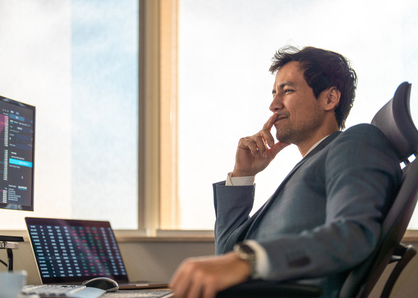 Focused mid adult Asian male stock broker in a modern office analyzes data on multiple screens, wearing a tailored suit and seated in a sleek chair.